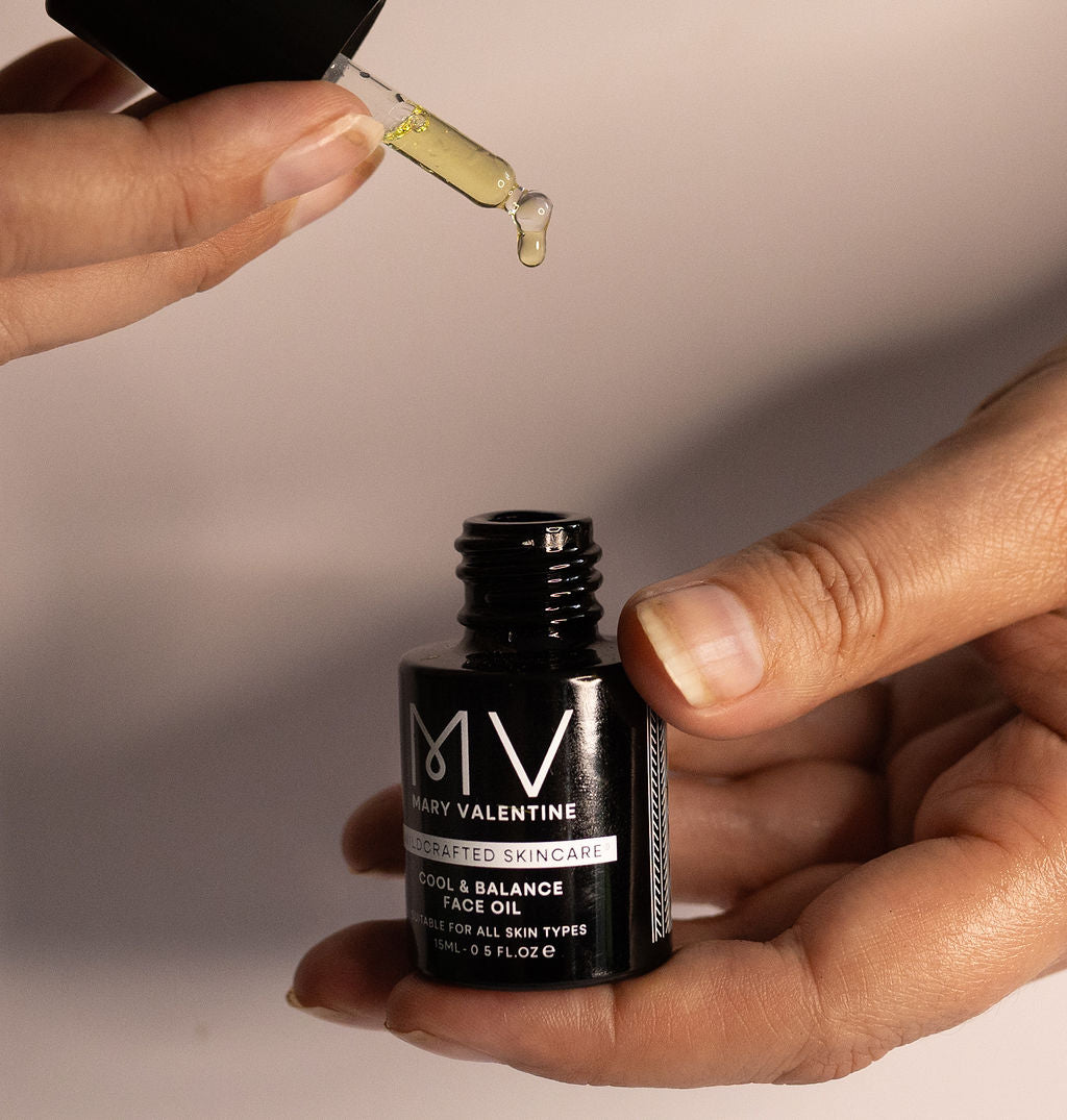 Hands holding an open bottle of Mary Valentine Wildcrafted Skincare Cool & Balance Face oil with a drop of oil coming from the pipette.